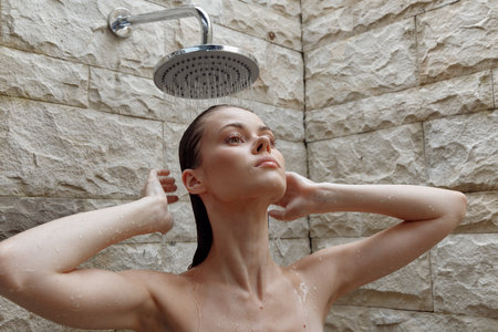young woman taking shower under rain showerhead with water droplets on skin against stone wall...