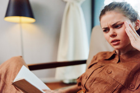 Young woman expressing frustration while reading a book in a cozy living room setting, wearing a...