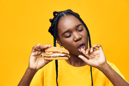 Happy Black woman enjoying a delicious burger against a vibrant yellow background, showcasing a fun and playful moment of indulgence in foodの写真素材