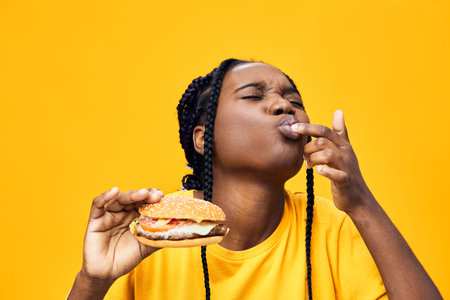 Hungry woman enjoying a delicious burger against a vibrant yellow background, showcasing joy and satisfaction in her expression, perfect for food related contentの写真素材