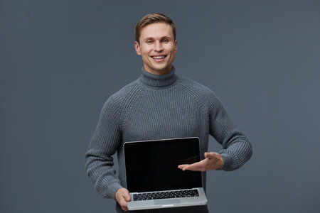 Confident smiling young Caucasian man in gray sweater holding laptop computer on plain solid blue background studio portrait. People lifestyle conceptの写真素材