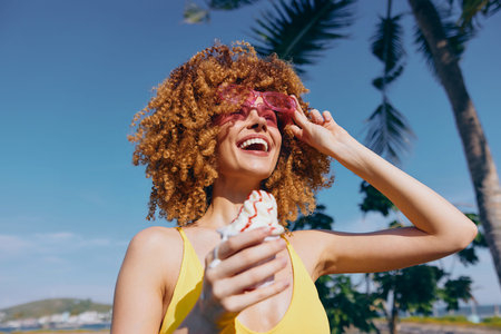 Happy woman with curly hair enjoying ice cream by the beach on a sunny day, wearing pink sunglasses and a yellow swimsuitの写真素材