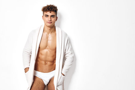 Confident young man with curly hair wearing a white robe, posing shirtless against a clean white background. Fitness and lifestyle concept.の写真素材