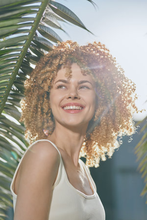 smiling woman with curly hair enjoying sunny day outdoors, green palm leaves background, natural...