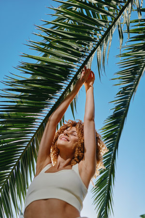 smiling woman with curly hair reaches up to green palm leaves under clear blue sky, wearing casual white top, sunlight highlighting joyful summer mood.の写真素材