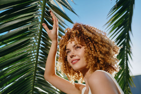 smiling woman with curly hair enjoying sunlight under tropical palm leaves against clear blue sky in summer atmosphereの写真素材