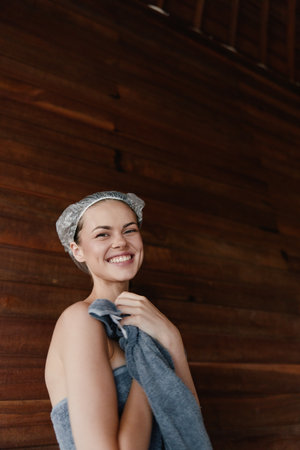 smiling woman with towel wearing shower cap stands against dark wooden wall, showing fresh clean skin and natural beauty after bath, wellness and relaxation conceptの写真素材
