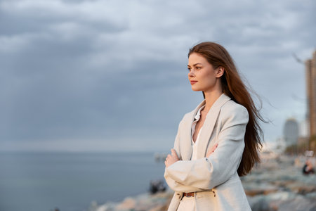 Confident businesswoman in a stylish blazer gazing at the horizon by the coastal cityscape, embodying ambition and strength against a dramatic sky.の写真素材