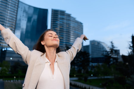 Confident businesswoman with arms outstretched, enjoying life in a modern urban setting, dressed in light professional attire, exuding joy and empowerment.の写真素材