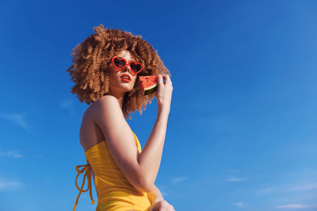 Young woman enjoying summer with a slice of watermelon against a clear blue skyの写真素材