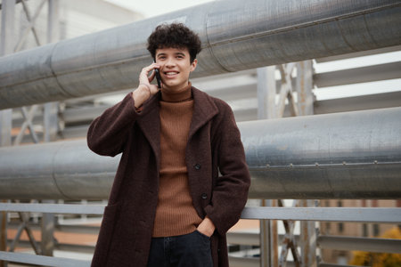 Fashionable man in brown coat talking on cell phone on street in front of metal pipe railing and...