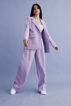 Young woman wearing a lilac pantsuit and white sneakers stands confidently against a soft purple background, showcasing modern fashion and relaxed style.の写真素材