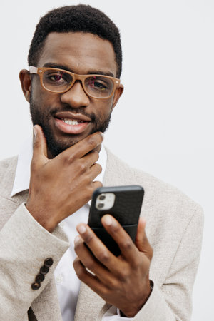 Stylish young African American man wearing glasses and a beige blazer holding a smartphone, thinking and smiling, isolated on a plain white background. People lifestyle conceptの写真素材