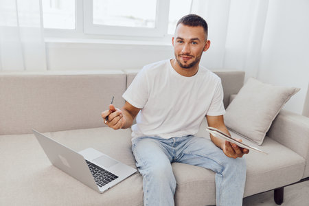Engaging man sitting on a couch, working on a laptop with a tablet in hand. Bright, modern interior with a cozy atmosphere. Perfect for remote work concepts.の写真素材