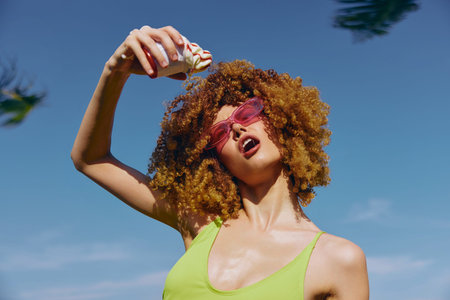 Young woman with curly hair in sunglasses enjoying an ice cream against a bright blue sky