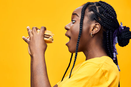Young Black woman with braids joyfully eating a burger against a vibrant yellow background, showcasing fun, youthfulness, and delicious fast foodの写真素材