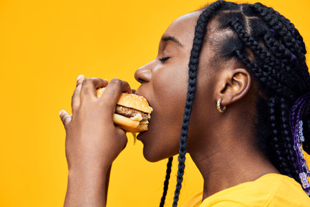 Young Black woman enjoying a hamburger with a bright yellow background, showcasing happiness and the joy of indulgent eating, perfect for food and lifestyle themesの写真素材