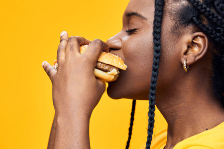 Young woman enjoying a delicious burger against a vibrant yellow background, showcasing her love for fast food and indulgenceの写真素材