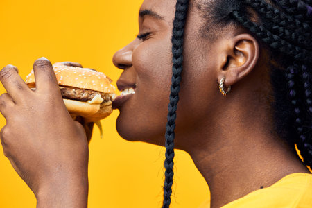 Smiling African woman enjoying a delicious hamburger, showcasing happiness against a vibrant yellow...