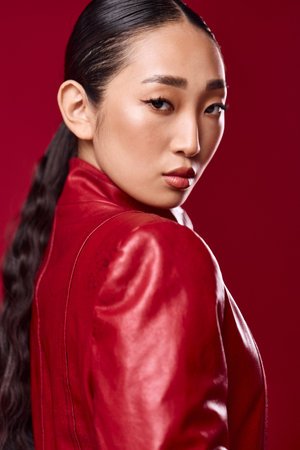 Fashionable asian woman in red leather jacket posing against red background with hand on hip and hair in ponytailの写真素材