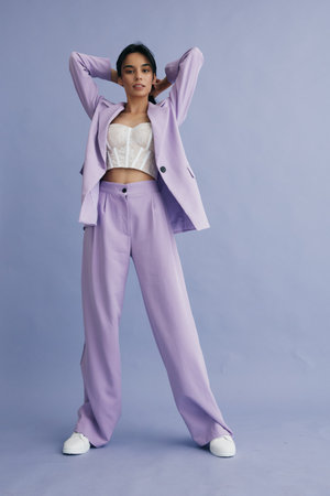stylish woman wearing lilac suit with white corset and sneakers posing confidently against soft purple background in modern fashion editorial styleの写真素材