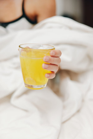 Woman holds glass of orange juice in bed with white comforter and sheetの写真素材