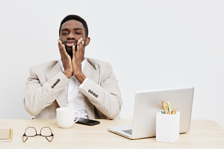 Frustrated young African American man in formal blazer sitting at desk with laptop, coffee mug, glasses, and smartphone, illustrating workplace stress and anxiety, indoor office environment, neutral background, modern business concept.の写真素材