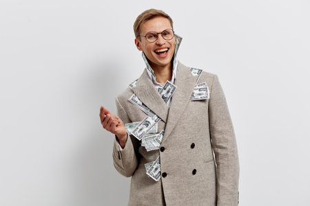 laughing young caucasian man wearing beige coat with money bills in pockets and joyful expression on...
