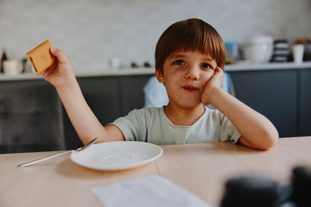 Thoughtful boy holding a piece of bread, sitting at the table. He looks curious and contemplative, with a focused expression in a cozy kitchen setting.の写真素材