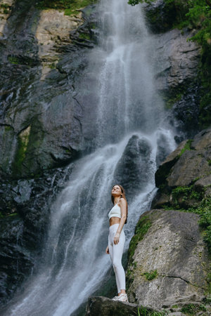 young woman standing near a tall waterfall in white activewear, surrounded by lush green forest and rocky cliffs, enjoying nature and serenityの写真素材