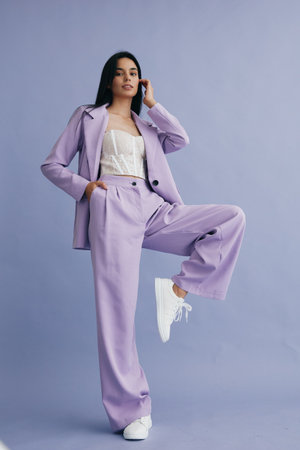 young woman in lilac suit posing confidently against plain purple background wearing white sneakers and lace top, stylish modern outfit, casual fashion lookの写真素材