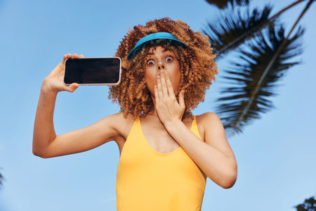 Surprised woman taking a selfie by the pool in a yellow swimsuit with a blue visor, tropical...