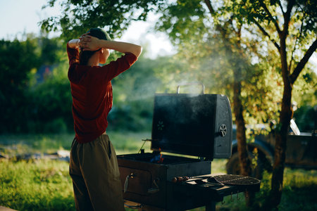 A young woman cooking on a barbecue grill outdoors in a green park, enjoying a sunny day with a relaxed expression, perfect for summer gatheringsの写真素材