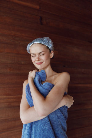 young woman wrapped in blue towel with shower cap enjoying spa relaxation, natural wooden background, peaceful expression, wellness and self-care conceptの写真素材