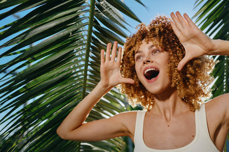 Smiling woman with curly hair enjoys bright sunlight under tropical palm leaves, wearing a white tank top and expressing joyful summer vibes outdoors.の写真素材