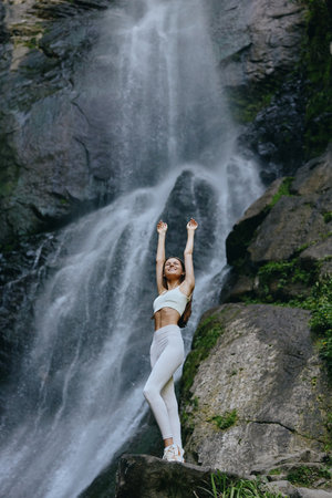 young woman stretching near waterfall in activewear, enjoying nature, wellness, and outdoor adventure, lush green rocks surrounding, serene mood.の写真素材