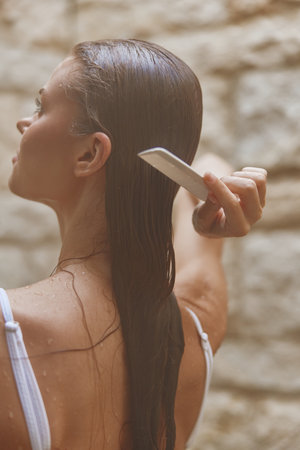 Woman combing wet long hair in a shower with natural stone wall background, showing self-care and relaxation in a serene bathroom setting.の写真素材