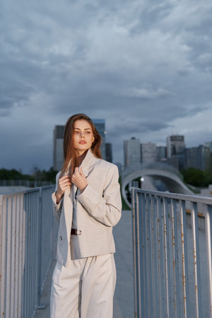 Woman in beige business suit standing on urban bridge with cloudy sky in background showcasing confident fashion style and modern city lifestyleの写真素材