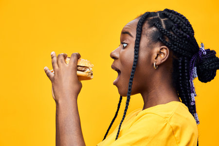Young Black woman excitedly holding a juicy hamburger against a vibrant yellow background, showcasing her love for fast food in a lively, playful atmosphereの写真素材