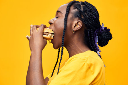 Young woman with braided hair enjoying a delicious burger against a vibrant yellow background, conveying joy and indulgence in fast foodの写真素材