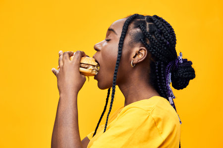 Young woman with braids enjoying a delicious burger against a vibrant yellow background, showcasing joy and a casual lifestyle in summer attireの写真素材