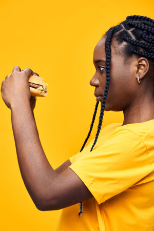 A young African woman in a yellow shirt thoughtfully holds a burger, showcasing healthy eating habits and vibrant emotions against a bright yellow backgroundの写真素材