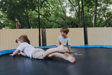 Two happy children playing on a trampoline, enjoying their carefree moments outdoors in a lively green setting with sunlight filtering through the trees.の写真素材