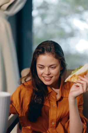 Young woman enjoying a healthy sandwich, smiling, dressed in a casual orange shirt, sitting in a cozy caf with soft natural lightの写真素材