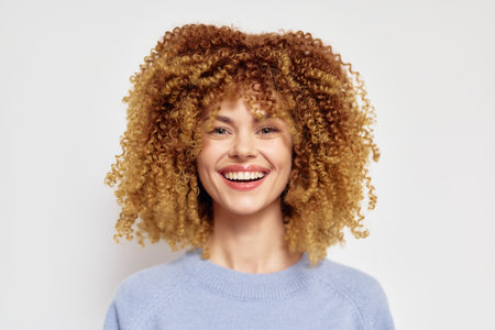 Smiling woman with voluminous curly hair, wearing a light blue sweater against a plain white background, showcasing natural beauty and confidence Her joyful expression radiates positivityの写真素材