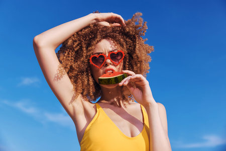 Young woman with curly hair in sunglasses holds a slice of watermelon against a bright blue sky Her playful pose conveys a fun summer vibeの写真素材