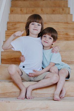 Two happy boys sitting on wooden stairs, joyfully embracing each other with smiles, showcasing friendship and childhood innocence in a warm light.の写真素材