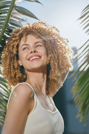 smiling woman with curly hair enjoying sunlight outdoors, bright joyful expression, tropical leaves...