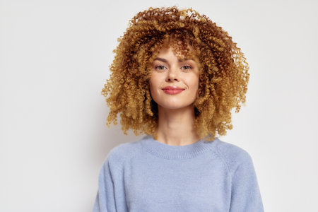 Beautiful woman with curly hair smiling in a light blue sweater, against a neutral white background, showcasing confidence and joy in a portrait styleの写真素材