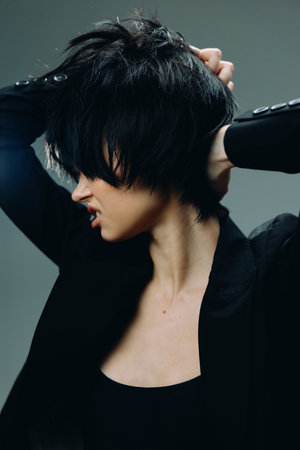 woman with short black hair in dark clothing pulling at her hair, expressing frustration and tension in a moody studio setting with muted gray backgroundの写真素材